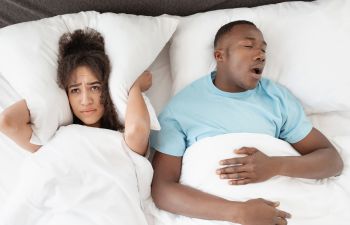 A loudly snoring man sleeping next to an annoyed woman covering her ears with a pillow.